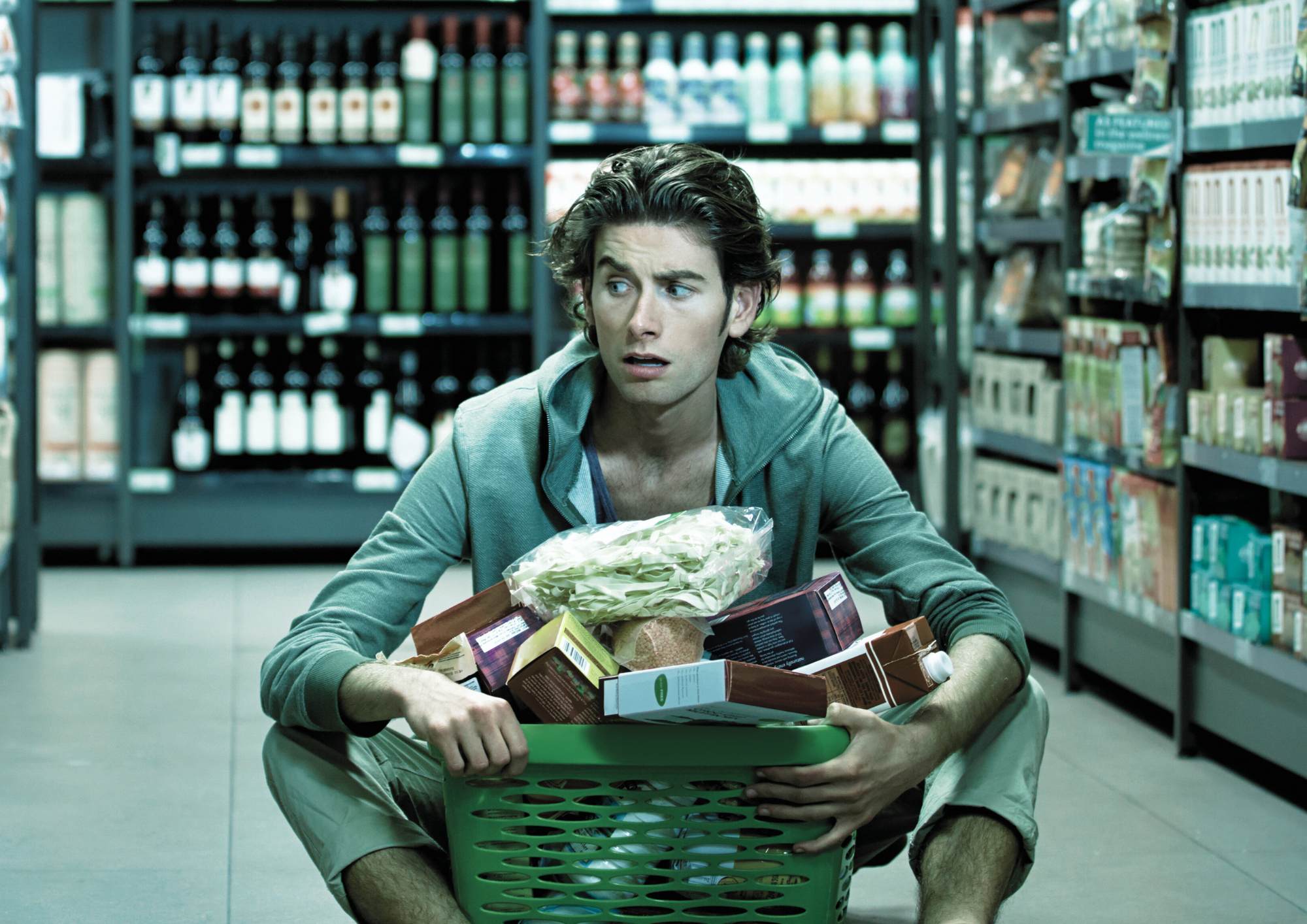 Young man in light green hoodie sitting in grocery store wine aisle, holding green shopping basket filled with food items, looking confused and overwhelmed by the wine selection on the shelves behind him